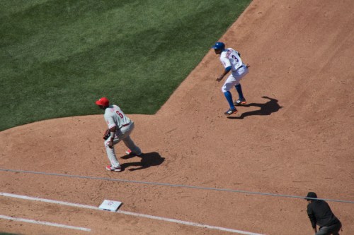 A baseball player on first base looks toward second base while another player runs toward him on the field.