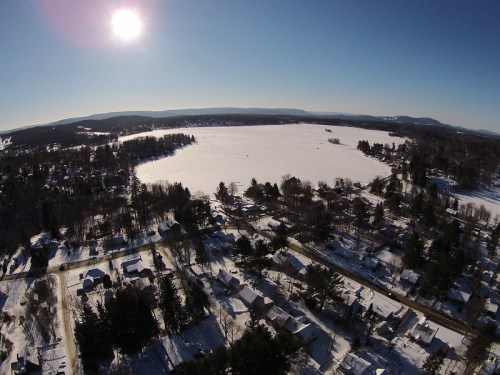 Aerial view of a snowy landscape with a frozen lake, surrounded by trees and houses under a clear blue sky.