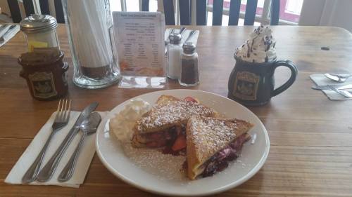 A plate of stuffed French toast with berries, whipped cream, and powdered sugar, set on a wooden table with utensils.