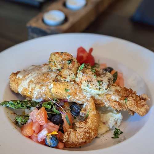 A plate of fried fish topped with shrimp, served with asparagus, tomatoes, blueberries, and a creamy sauce.
