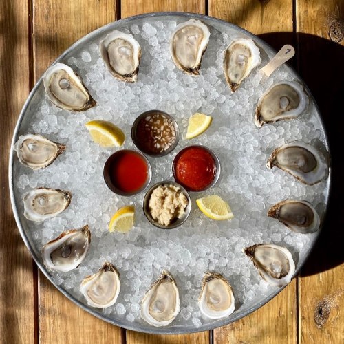 A circular platter of fresh oysters on ice, surrounded by lemon wedges and three dipping sauces.