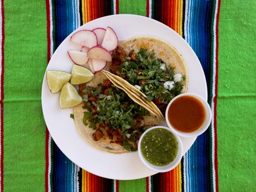 A plate of tacos topped with cilantro, served with radishes, lime wedges, and two sauces on a colorful striped tablecloth.