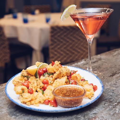 A plate of crispy calamari with cherry tomatoes and a dipping sauce, alongside a pink cocktail with a lime wedge.