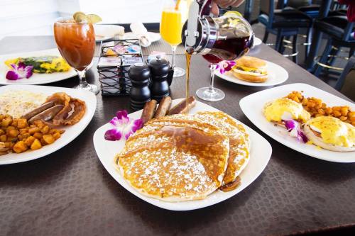 A table set with breakfast dishes: pancakes, sausages, eggs, and drinks, garnished with flowers. Syrup is being poured.