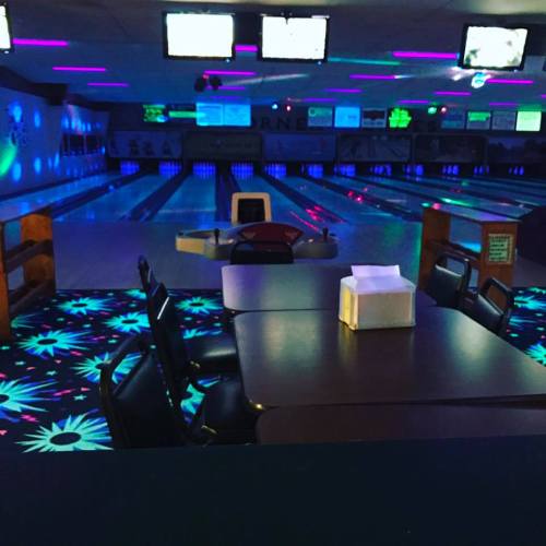 A dimly lit bowling alley with neon lights, empty lanes, and a table in the foreground.