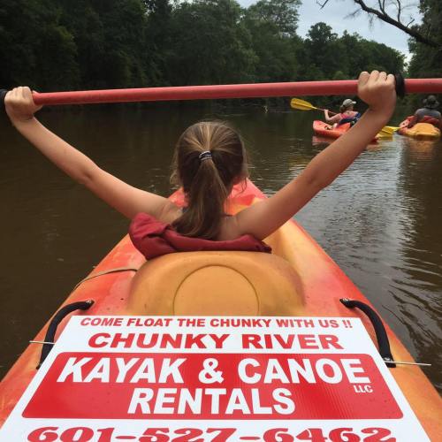 A person paddles a kayak on a river, with a sign for Chunky River Kayak and Canoe Rentals visible in the foreground.