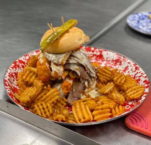A towering sandwich with layers of meat, topped with a pickle, served on a plate of waffle fries.
