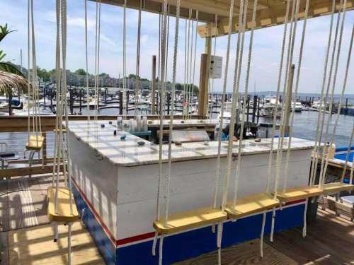 A unique bar with swings for seating, overlooking a marina filled with boats and a clear blue sky.