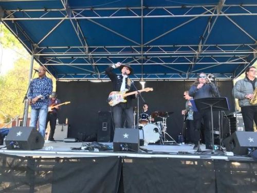 A band performs on stage with various instruments, including guitars and brass, under a blue canopy.