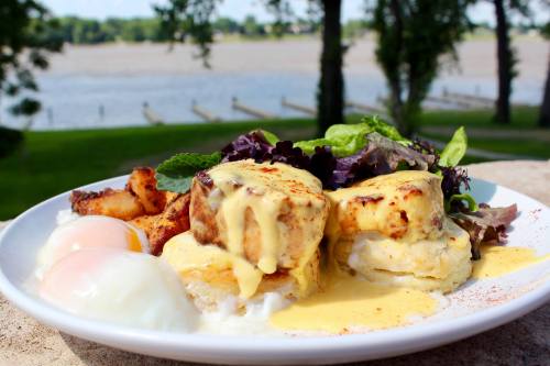 A plate of eggs Benedict with poached eggs, hollandaise sauce, and a side salad, set against a scenic river view.