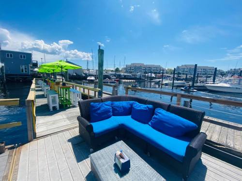 A cozy outdoor seating area with blue cushions on a dock, overlooking boats and a sunny marina.