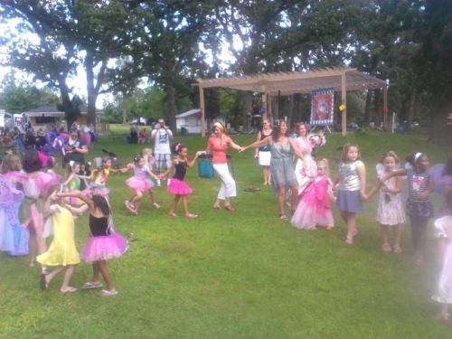 A lively outdoor scene with children in colorful costumes dancing and playing, surrounded by adults and trees.