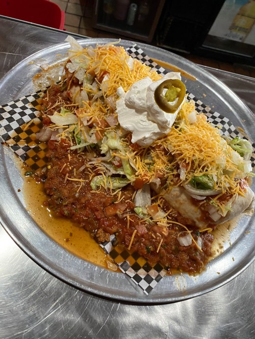 A large plate of burritos topped with cheese, lettuce, onions, sour cream, and jalapeños, served with chili sauce.