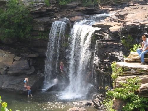 A waterfall cascades over rocks, with children playing in the water and a person sitting on the edge nearby.