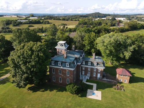Aerial view of a large historic brick house surrounded by trees and open fields under a blue sky with clouds.