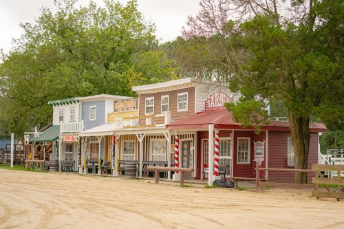 A row of colorful, old-fashioned storefronts in a western-style town, set against a backdrop of trees.
