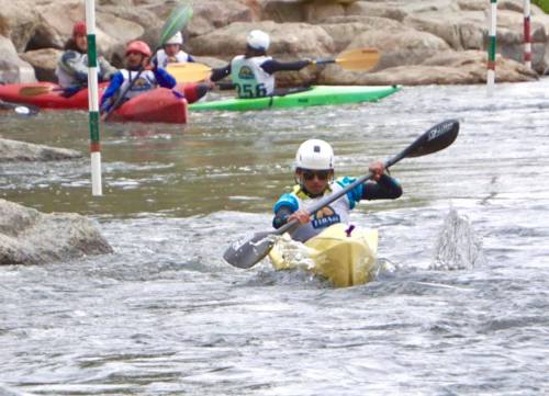 A kayaker navigates a river course with slalom poles, surrounded by other paddlers in colorful kayaks.