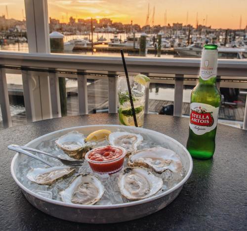 A platter of oysters on ice with cocktail sauce, a drink, and a beer, set against a sunset over a marina.