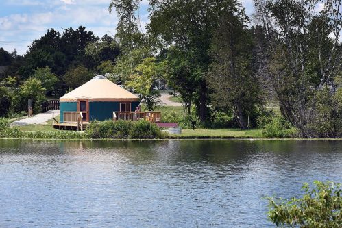 A yurt by a calm lake, surrounded by trees and greenery, with a wooden deck and a clear blue sky above.