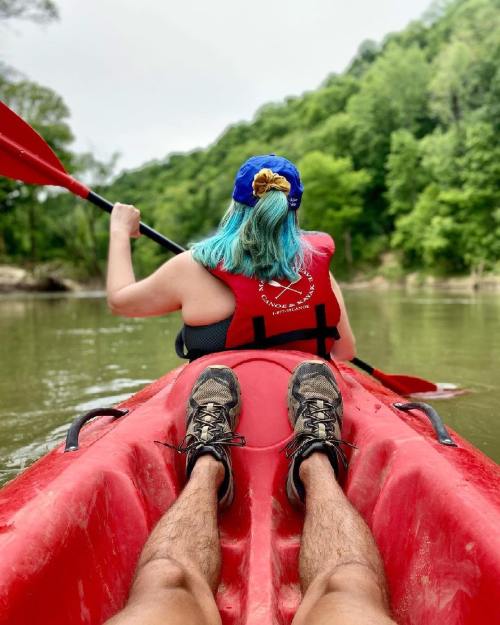 A person with blue hair paddles a red kayak on a calm river, surrounded by green trees.