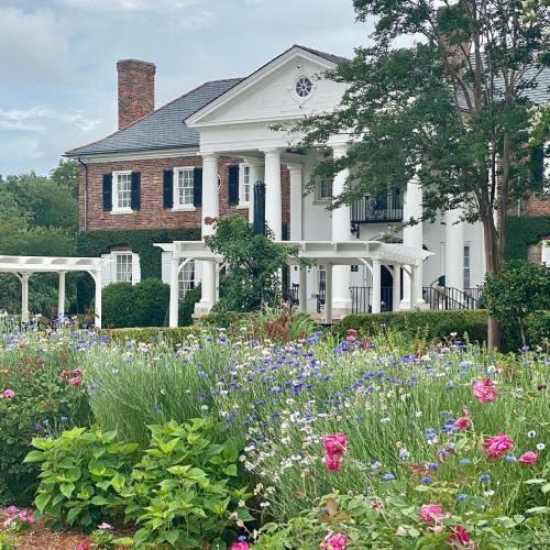 A charming brick house with white columns, surrounded by colorful flowers and lush greenery.