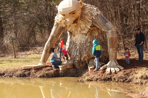 A large wooden sculpture of a troll sits by a pond, surrounded by children and adults exploring the area.