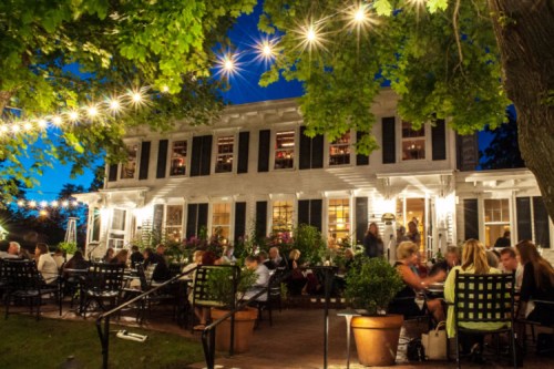 A lively outdoor dining scene at night, featuring a charming restaurant with string lights and guests enjoying their meals.