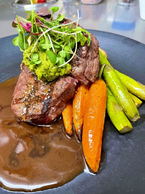 A beautifully plated steak topped with greens, served with carrots and leeks, drizzled with sauce on a black plate.