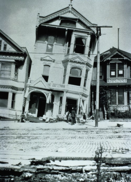 A tilted house after an earthquake, with damaged buildings and people in the foreground on a cobblestone street.