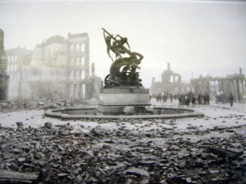 A damaged statue stands in rubble, surrounded by ruins and people walking in a desolate, war-torn landscape.