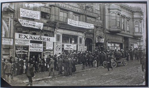 Historic street scene with crowds and horse-drawn carriages outside businesses, featuring signs for printing and services.