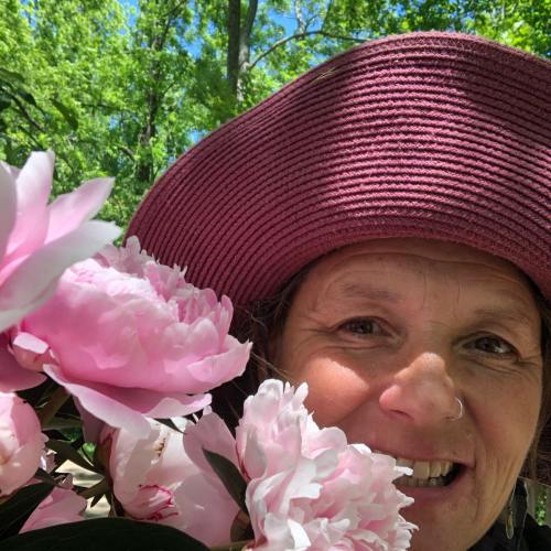 A woman smiles while surrounded by pink flowers, wearing a wide-brimmed hat in a lush green setting.