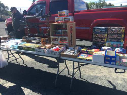 A vendor at a flea market displays various toys and collectibles on a table beside a red pickup truck.