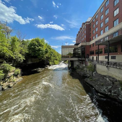 A river flows beside a hotel, surrounded by lush greenery and a clear blue sky with scattered clouds.
