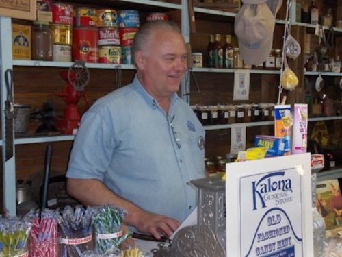 A smiling man stands behind a counter at Kalona General Store, surrounded by colorful candy and vintage items.