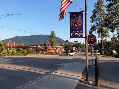 A street view of St. Regis with a welcome banner, gas stations, and mountains in the background under a clear blue sky.