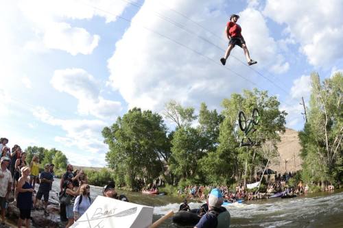 A person zip-lining over a river while spectators watch from the shore, with trees and clouds in the background.