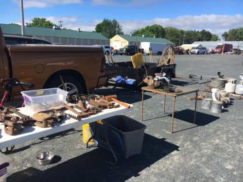 A flea market scene with various tools and items displayed on tables beside a parked truck.