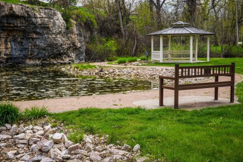 A serene park scene featuring a gazebo by a pond, with a wooden bench and rocky shoreline surrounded by greenery.