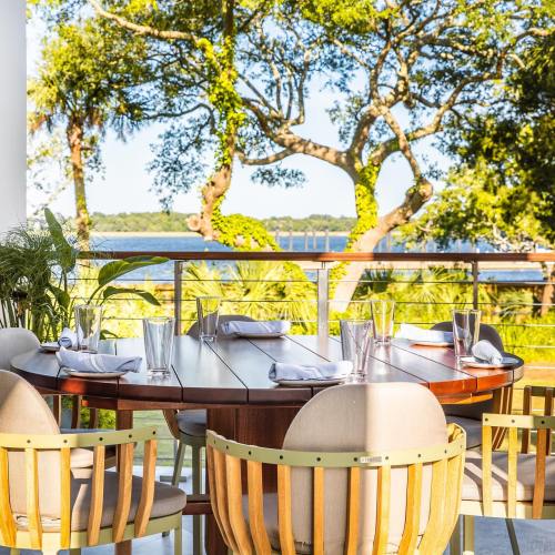 A scenic outdoor dining area with a round wooden table, chairs, and a view of trees and water in the background.