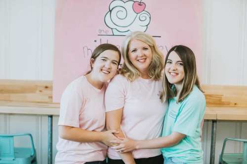 Three women smiling together in a cozy setting, wearing matching pastel shirts, with a pink wall in the background.