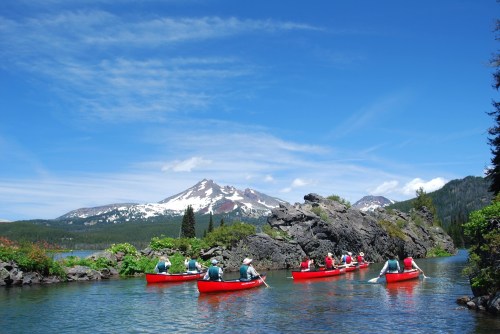 A group of people in red canoes paddling on a serene lake with mountains and blue skies in the background.
