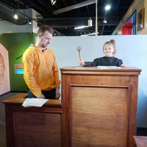 A young girl in a judge's robe holds a gavel, while a man in an orange hoodie stands beside her at a wooden podium.
