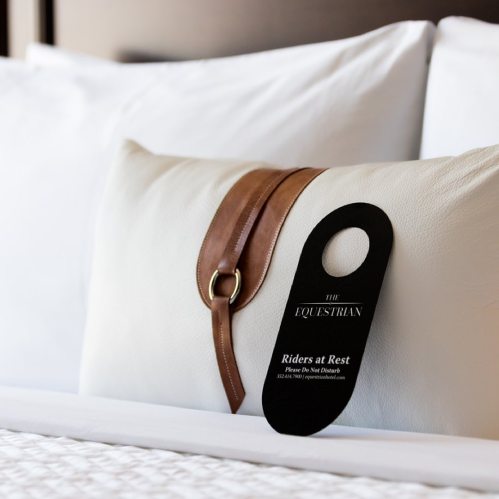 A decorative pillow with a leather strap and a tag reading "Riders at Rest" on a neatly made bed.