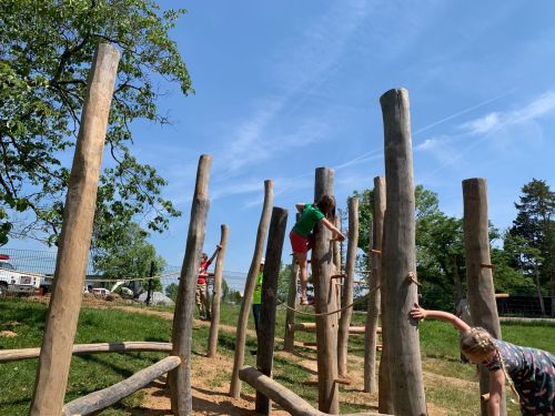 Children play on a wooden climbing structure in a sunny outdoor park, surrounded by trees and blue skies.