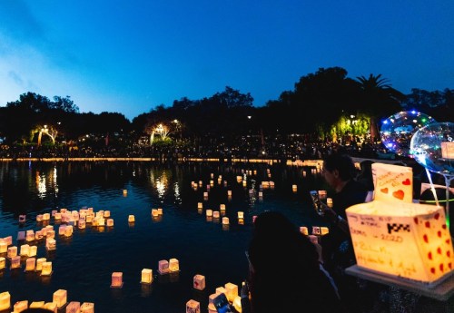 A serene evening scene with glowing lanterns floating on water, surrounded by trees and a crowd enjoying the view.