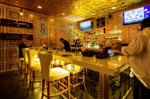 A stylish bar area with golden decor, featuring patrons, a bartender, and a well-stocked bar in a vibrant setting.