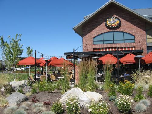 A restaurant exterior with red umbrellas, outdoor seating, and landscaped greenery under a clear blue sky.