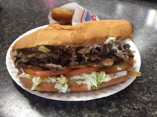 A loaded sandwich with sliced beef, onions, lettuce, and tomatoes on a bun, served with a side of fries.