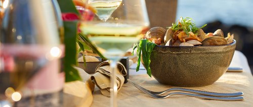 A close-up of a seafood dish in a bowl, with a glass of white wine and a scenic outdoor setting in the background.